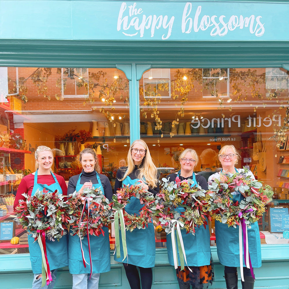 Five women holding floral wreaths in front of a store named 'the happy blossoms' they made at the happy blossoms flower school