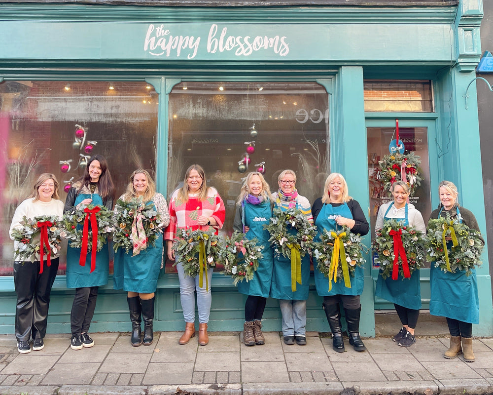 Group of women standing in front of a store named 'The Happy Blossoms' holding floral wreaths they made at the happy blossoms flower school